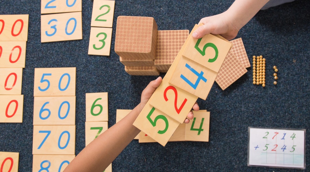 Two hands - one holding two number tiles and the other holding two number tiles to make a 4 digit number. The students are using Montessori materials to do their work.