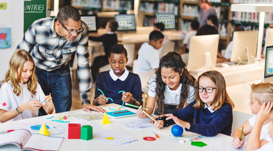 A picture of a make teacher observing a group of students working with 3D shapes at a table. 