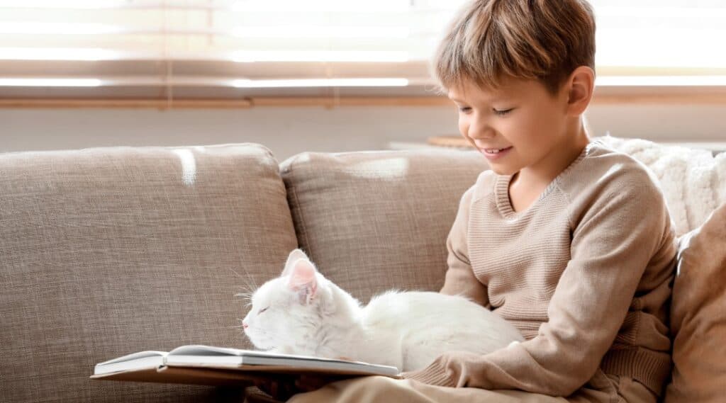 A 10 year old boy and his white cat sitting on a couch enjoying a cat book for kids.