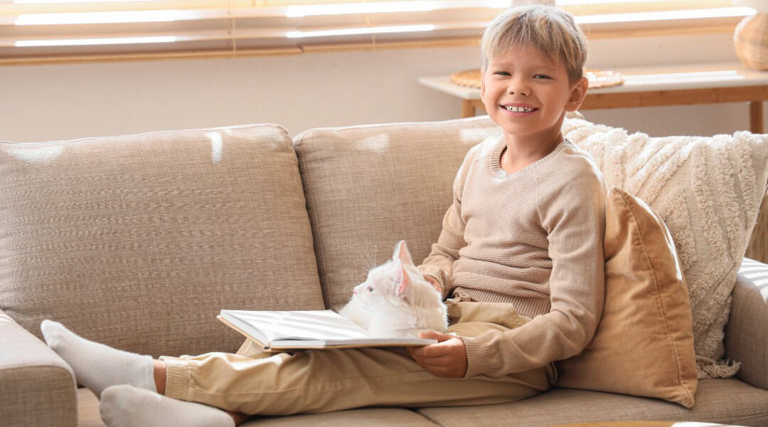 A 10 year old boy and his white cat sitting on a couch reading a cat book for kids.