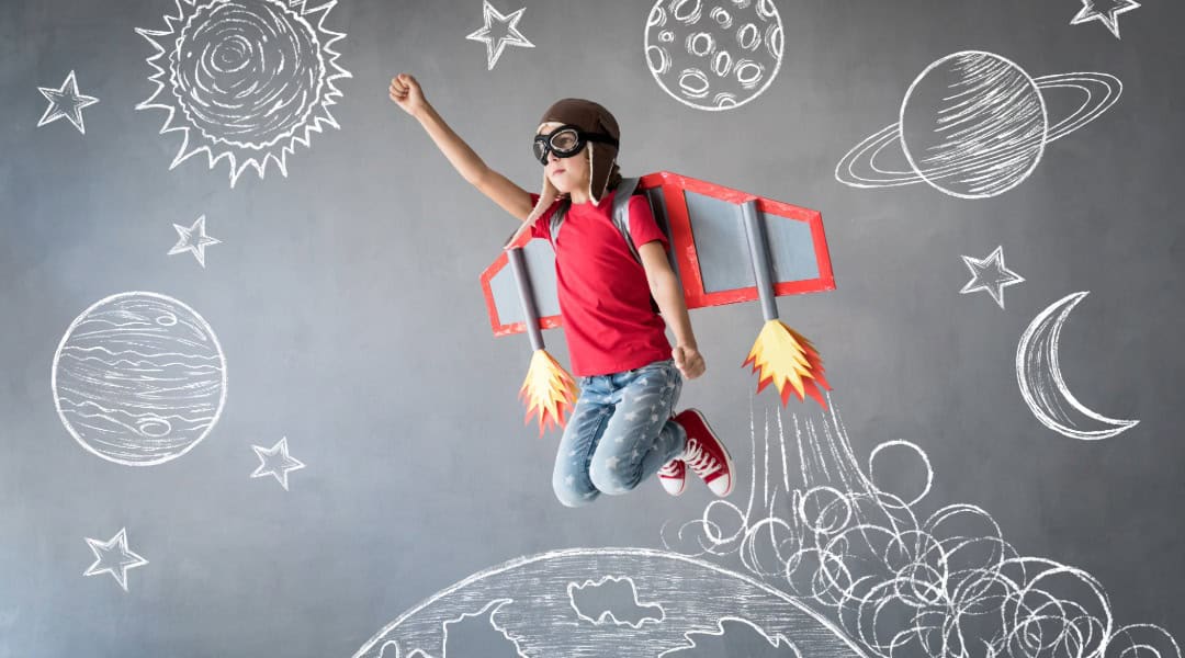 An elementary student with a cardboard rocket attached to their back and up against a backdrop that has space elements drawn in chalk.