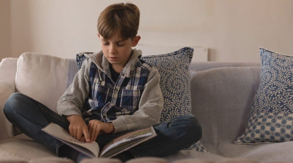 A ten year old boy sitting on a couch reading a picture book.
