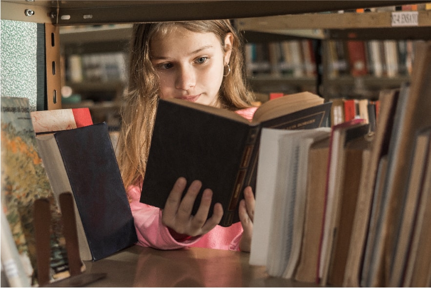 An 8 year old girl is scene through the book stack at a library. She has a book with a brown cover in her hands and she is reading it. She is choosing a book to use for one of our summer reading activities for elementary students.