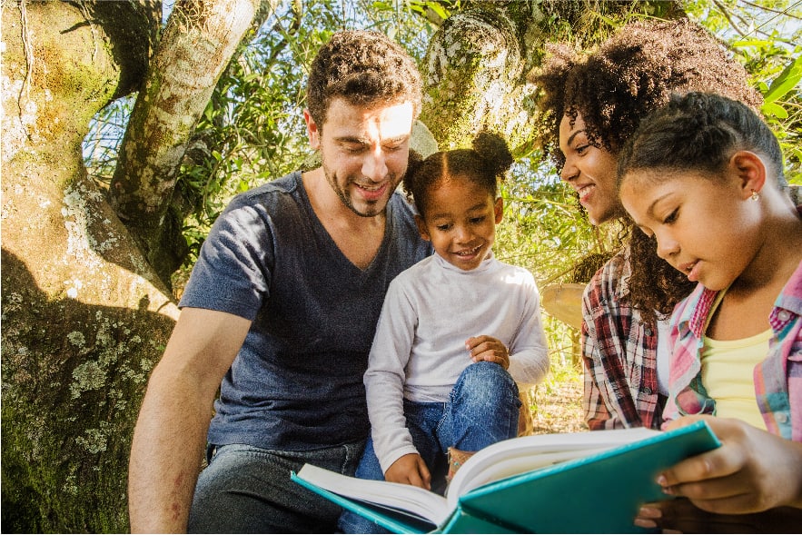 A dad, mom, and  their two lower elementary-aged daughters are sitting out in nature and have a book opened in front of them. They are reading a story together.