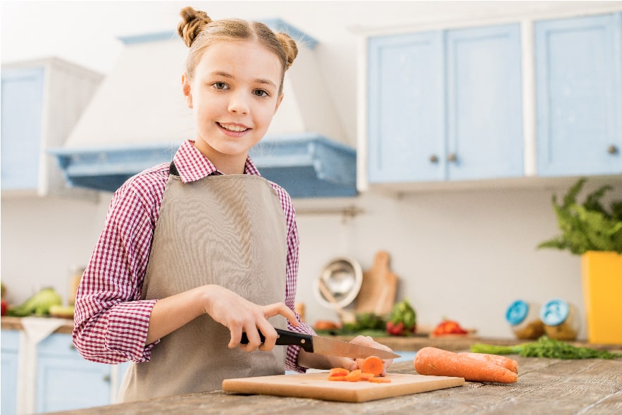 A picture of a young elementary-aged girl in the kitchen with an apron on. She has carrots on a cutting board. It is her turn to prepare the snack for the family book club meeting, an excellent summer reading activity!
