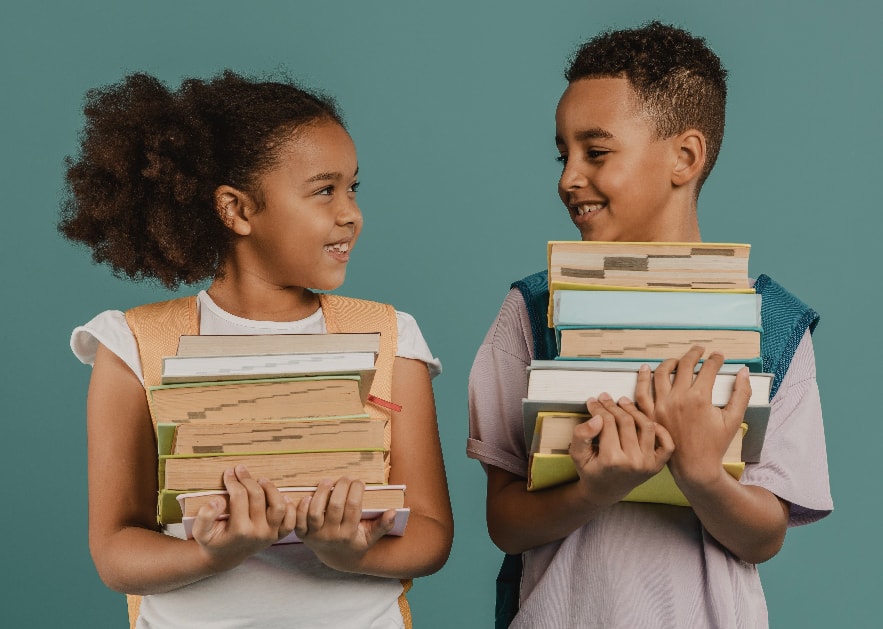 A picture of a boy and a girl both holding a stack of 5 books in their arms. They're going to donate these old books to the book drive. Another wonderful summer reading activity elementary students will benefit from.
