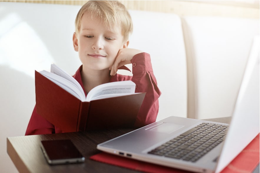 A picture of a 10 year old boy holding and reading a book while he is in front of his computer. There is also a phone on his desk. He is taking part in a virtual reading club.