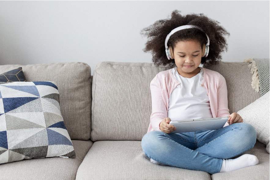 A 7 year old girl is sitting on a coach with headphones on. She is holding a tablet and smiling because she is listening to an audiobook. Listening to audiobooks is just one of many awesome summer reading activities elementary kids will love!