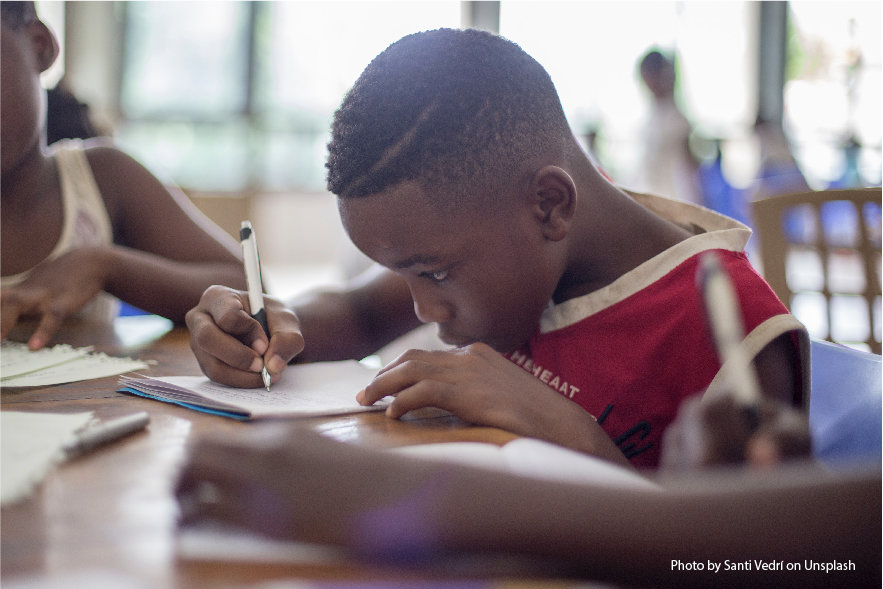 An image of a 9 year old boy sitting at a table with a pen in his right hand. He is focused on writing a letter. There is a girl in the background at the table with him and blurred children in the distant background. This is a classroom or library setting.