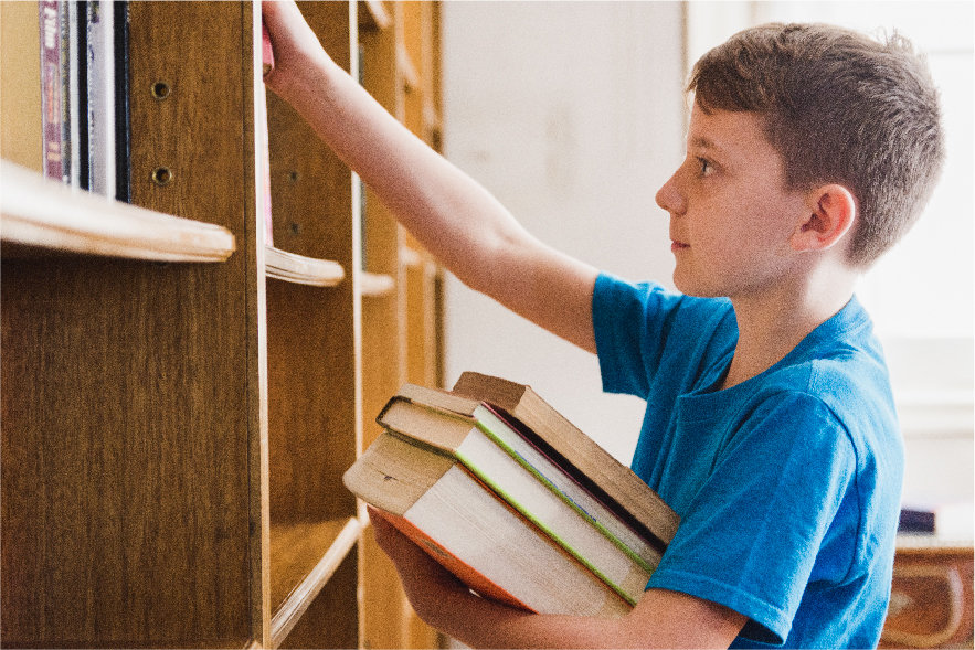 A picture of a young boy holding a stack of 3 books in his left arm and reaching into a book shelf with his right hand.