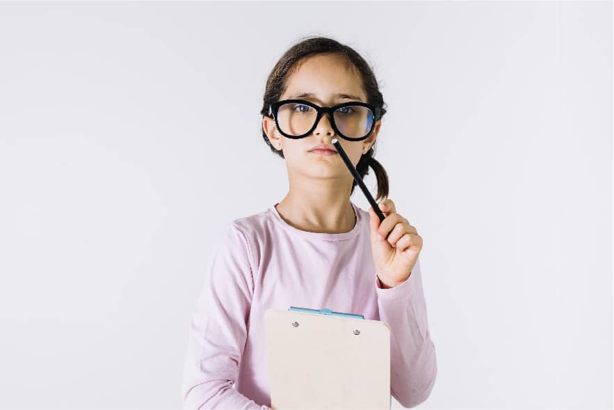An 8 year old girl is pictured wearing black rimmed glasses and a pink long sleeve shirt. She is pointing a pencil toward the camera with her left arm and holding a clipboard with her right arm.