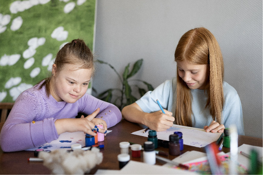 Two elementary-aged girls are sitting at a table that is covered with arts and crafts material. They are going to do some creative artwork related to something they read.