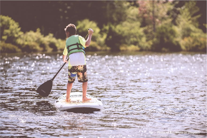 A picture of a young boy on a paddle board, standing on the board holding a black paddle and wearing a green lifejacket. This boy put paddle boarding on his bucket list and made it happen!