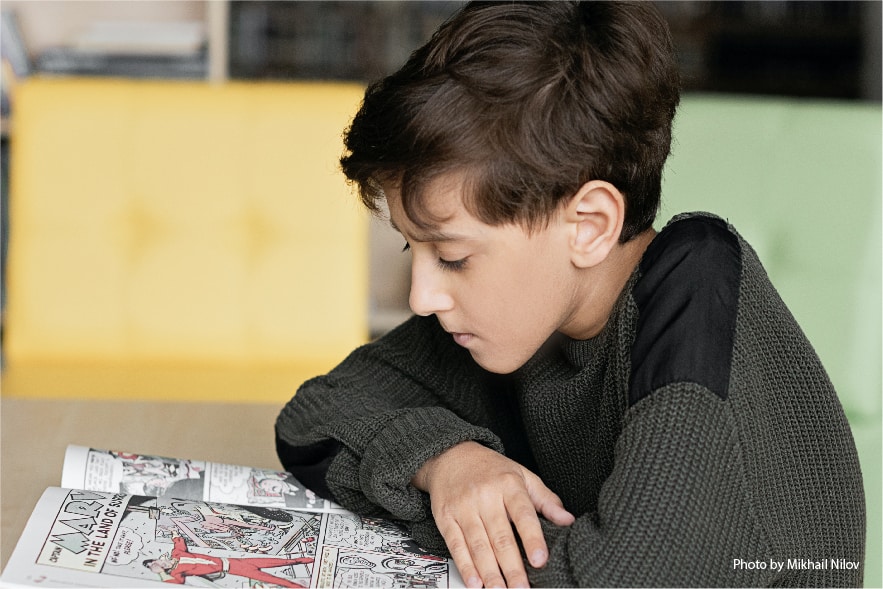 A picture of a 10 year old boy sitting at a table reading a comic book. 