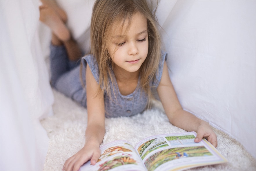 A picture of a young girl lying on a soft white carpet with a white canopy above her. She has a picture book in front of her and she's reading in a cozy little space. Reading in a fun location is just one of many awesome summer reading activities elementary kids will love!