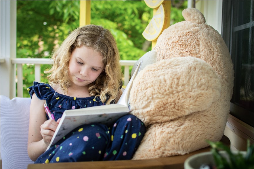 A picture of an 8 year old girl sitting on a couch with a big brown stuffy. She is writing in her reading log to keep track of her reading progress and practice. Reading logs are one of many awesome summer reading activities elementary kids will love!