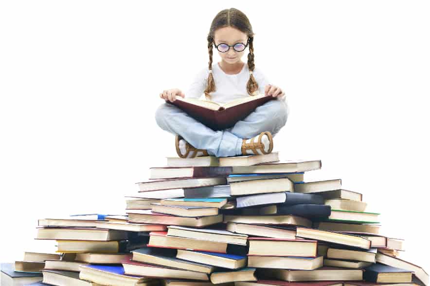 A picture of a 7 year old girl sitting on top of a pyramid-shaped stack of books. She is sitting cross-legged and reading a book.