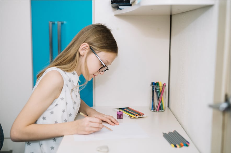 A picture of a 9 year old girl sitting at a desk drawing with a pencil on paper. She is making her own personalized bookmark, which is a great summer reading activity for elementary kids.