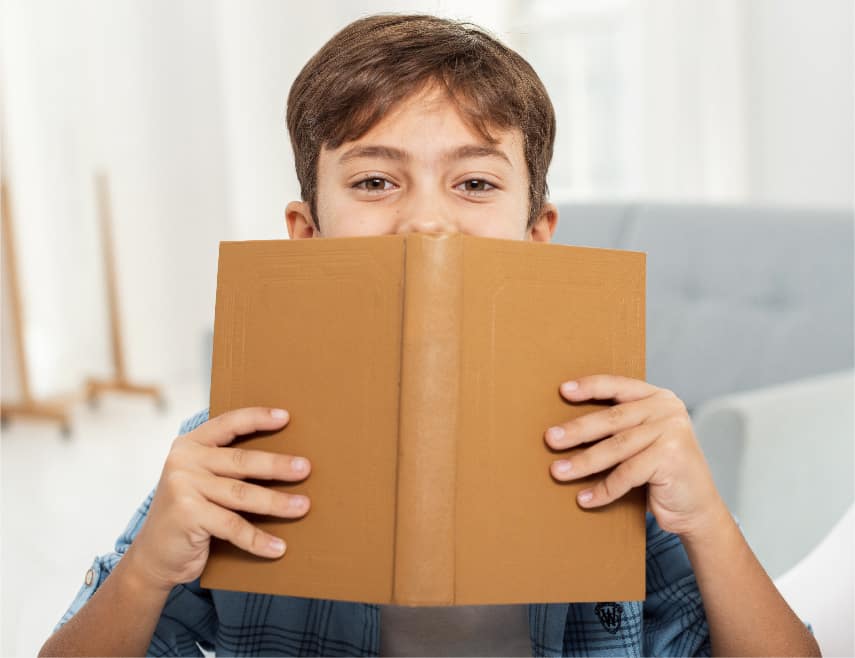A picture of a 9 year old boy holding a brown covered book up to his face covering his mouth.