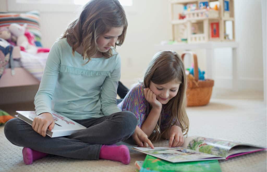 Two elementary aged girls working together. One girl is laying on her stomach on the floor looking at a picture book. The other girl is sitting cross-legged beside her friend. They are doing some classroom vocabulary activities.