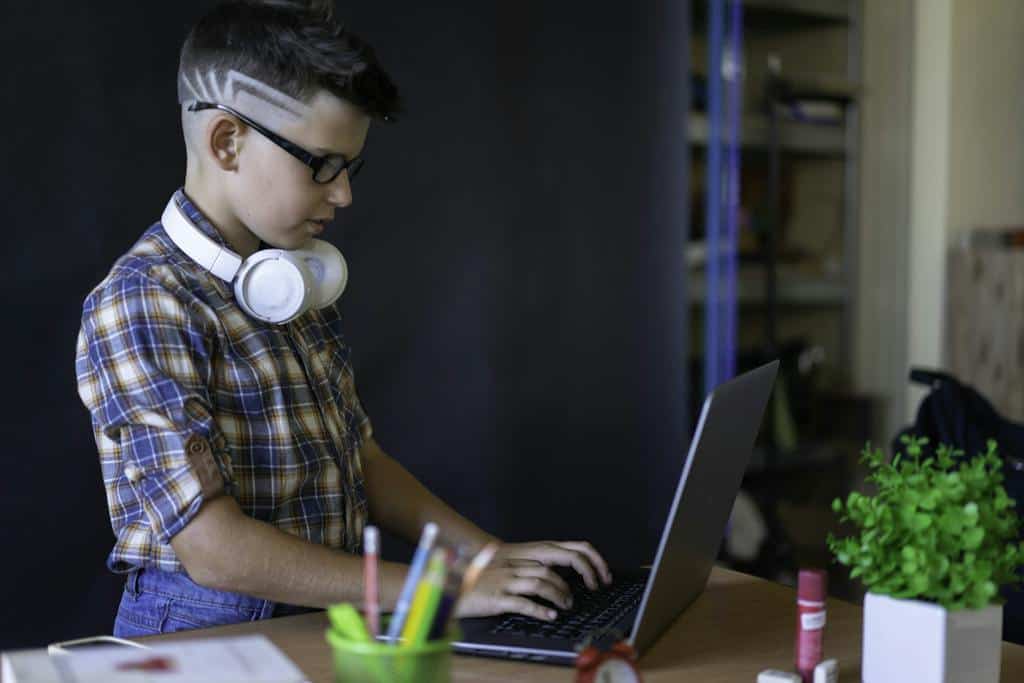A Boy Using a Laptop with headphones around his neck. He is typing information about the audiobook he just listened to.