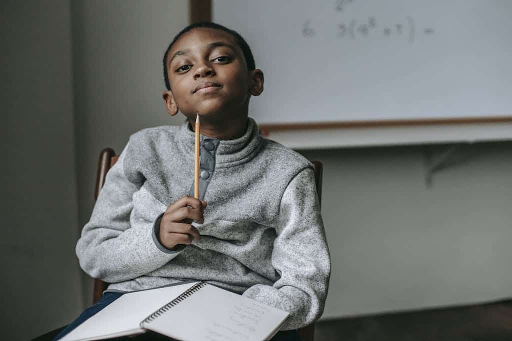 Cute little black boy wearing fleece sweater sitting on chair with pencil under chin brainstorming book report ideas for a 5th grade assignment.