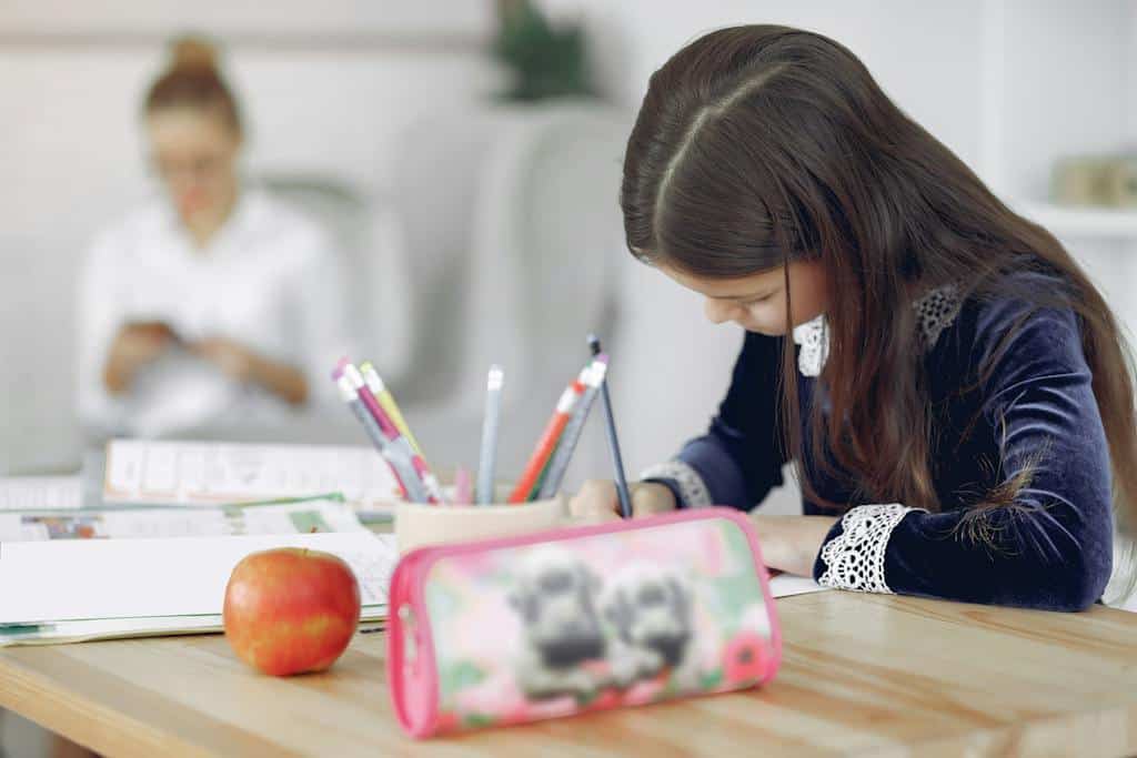 Focused young girl in school uniform sitting at table with stationery and apple while writing in a notebook, taking notes after watching some parts of speech videos.