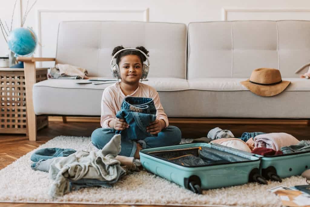 Girl in Long Sleeve Shirt Sitting on the Floor folding clothes to put in a suitcase. She is wearing headphones and listening to an audiobook.
