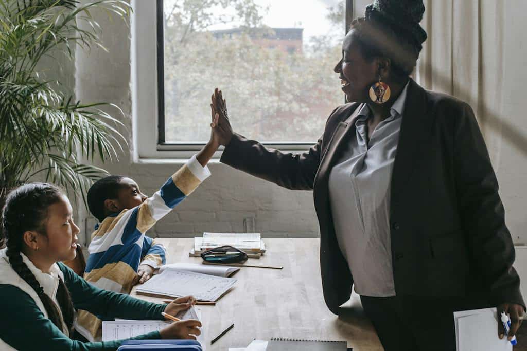Side view of smiling black boy giving high five to teacher while sitting with diverse classmate girl at desk in classroom. They are studying the parts of speech.