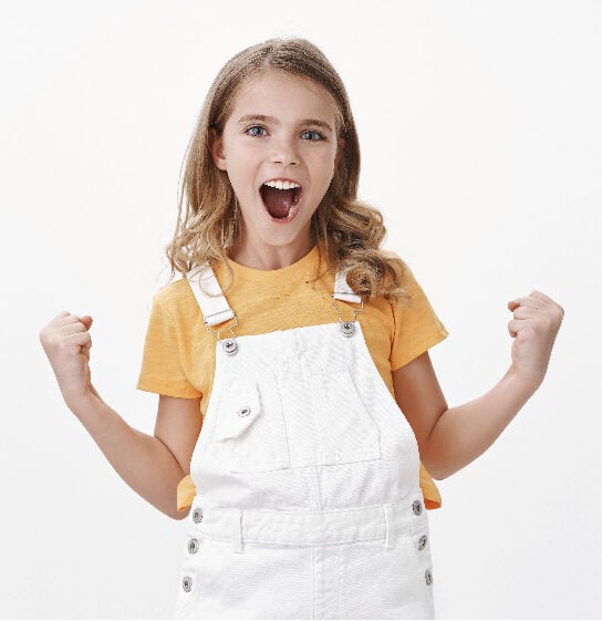 An image of a 9 year old girl wearing white overalls and a yellow t-shirt looking very happy, proud even.