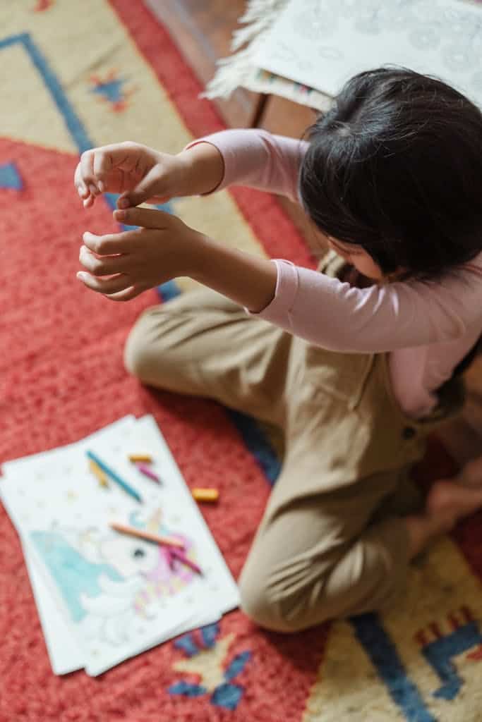 From above of full body anonymous female child in casual outfit sitting on carpet with drawings and crayons while dropping stationary from height. She's ready for some creative spring break activities for kids.
