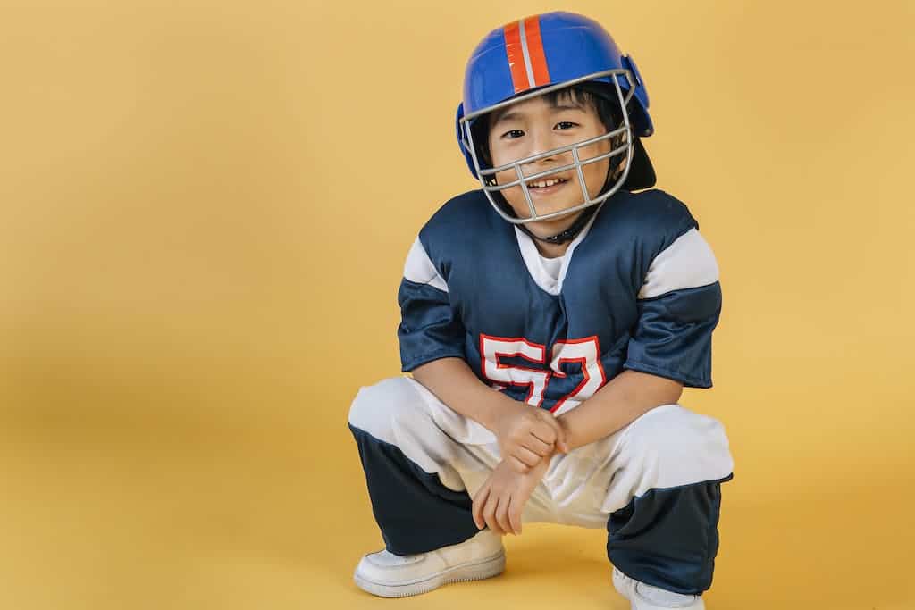 Positive Asian boy wearing protective helmet and sportswear of American football player in yellow studio