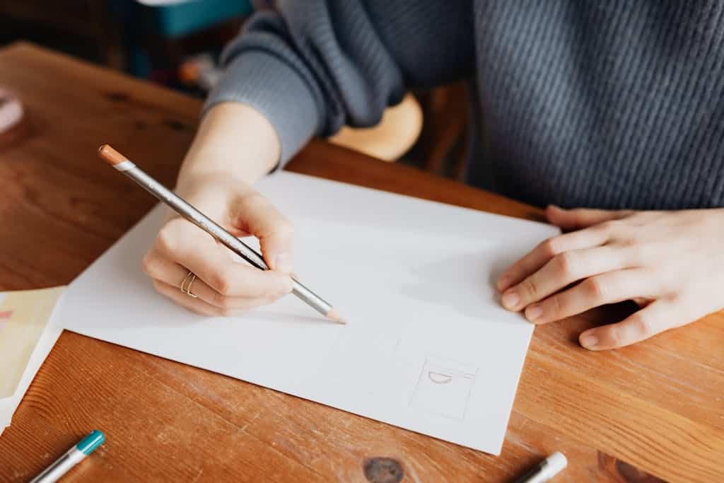 A grade 6 female student in gray sweater sitting at shabby table and holding a pencil to paper as she write about the answer she got for her rebus puzzle