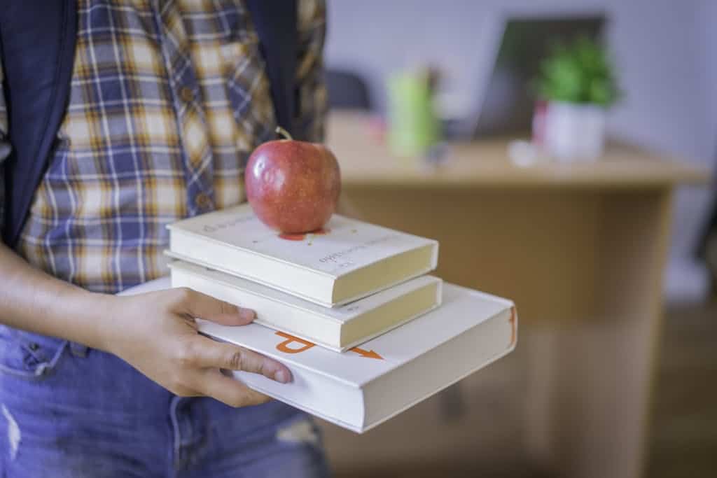 Child Holding a Stack of Books with an Apple on Top getting ready for a book tasting which is a great summer reading activity for kids.