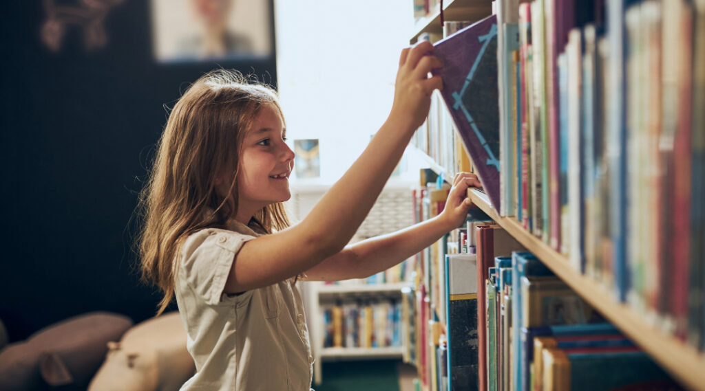 8 year old girl in library pulling a book off the shelf while smiling