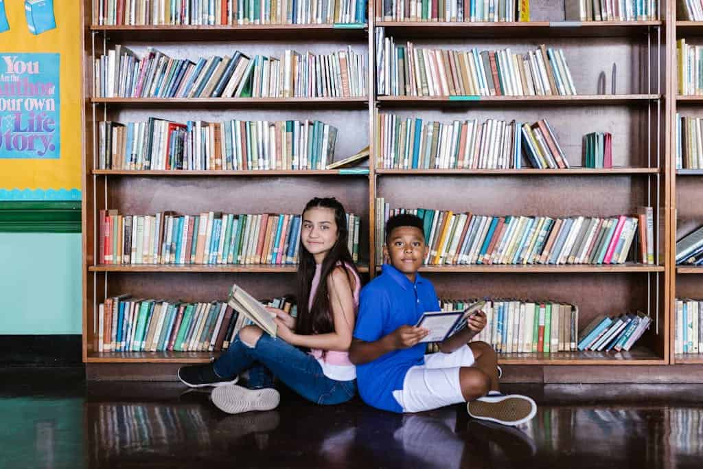 A Boy and Girl Sitting on the Floor in front of library book stacks While Holding Books. They are working together as a team to complete a library scavenger hunt.