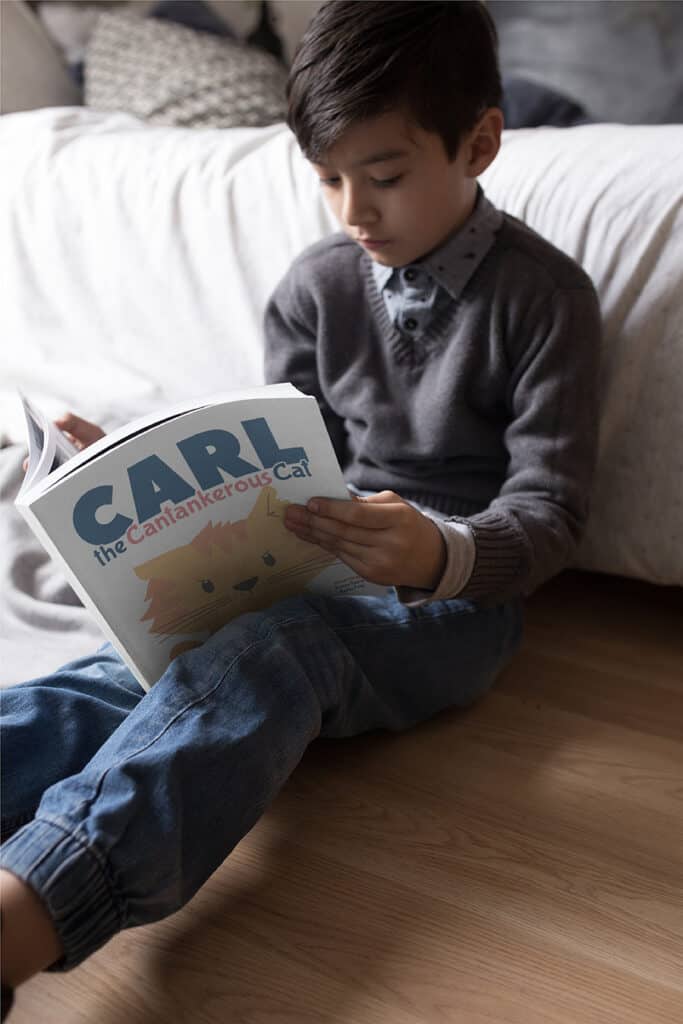 An 8 year old boy sitting on the floor reading Carl the Cantankerous Cat, which is one of the best picture books to teach vocabulary.