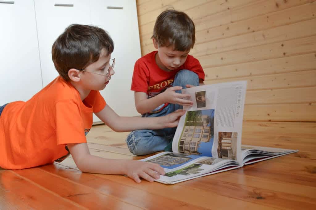 Two curious, smart siblings in casual clothes sitting on wooden floor and reading interesting magazine.