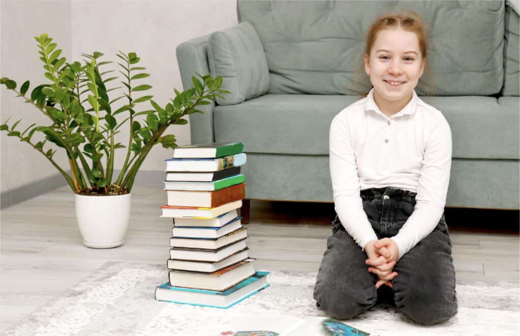 A 10 year old girl sitting on her knees on the carpet beside a stack of book recommendations she got from doing a scavenger hunt.