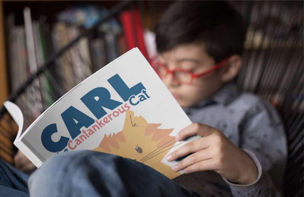 An image of an elementary-aged boy in red glasses sitting reading the picture book Carl the Cantankerous Cat.