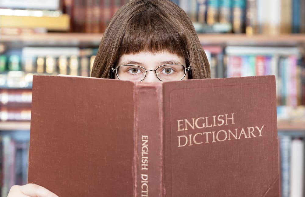 A picture of an elementary aged girl holding a dictionary up covering most of her face. You can see her glasses and nose.
