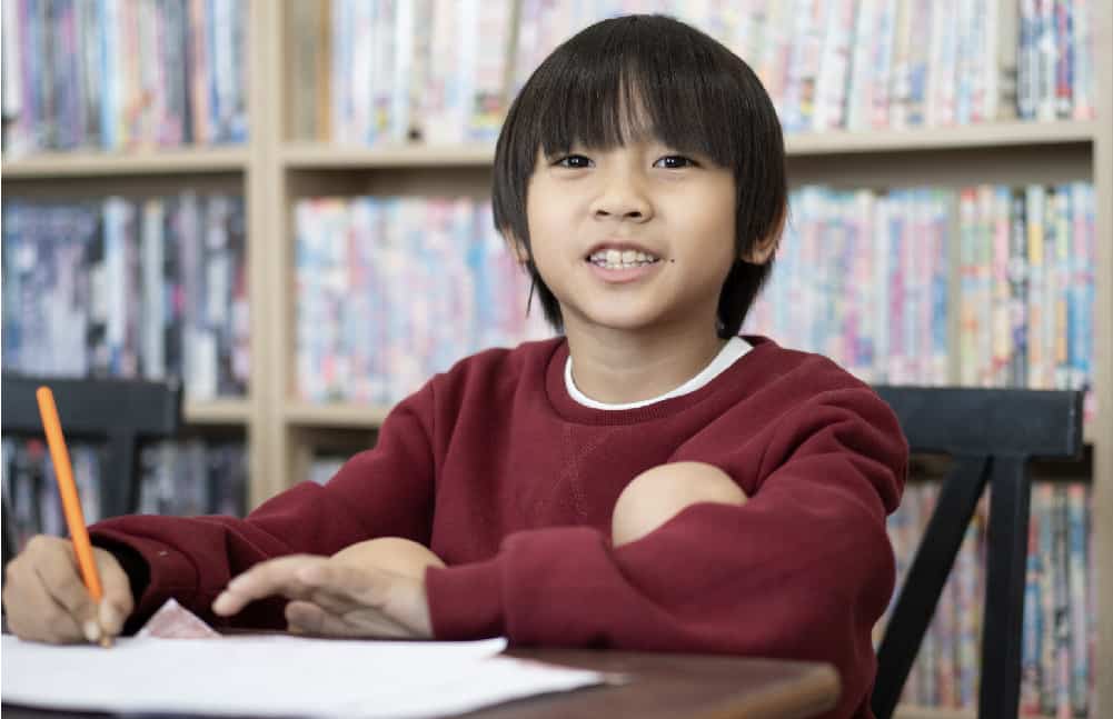 A picture of an elementary-aged boy sitting at a table in the library writing a research paper from a topic he discovered while doing a scavenger hunt.