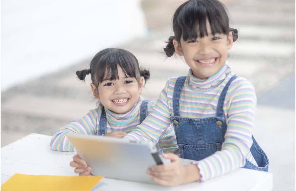 An elementary-aged girl and her younger sister are both dressed in jean overalls with a stripped long sleeve shirt. They are getting ready to do a library scavenger hunt.