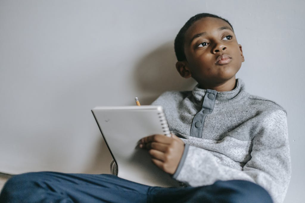 Little thoughtful black kid sitting on floor with notebook and pencil looking away against white wall as he ponders the short quotes for students he has read.