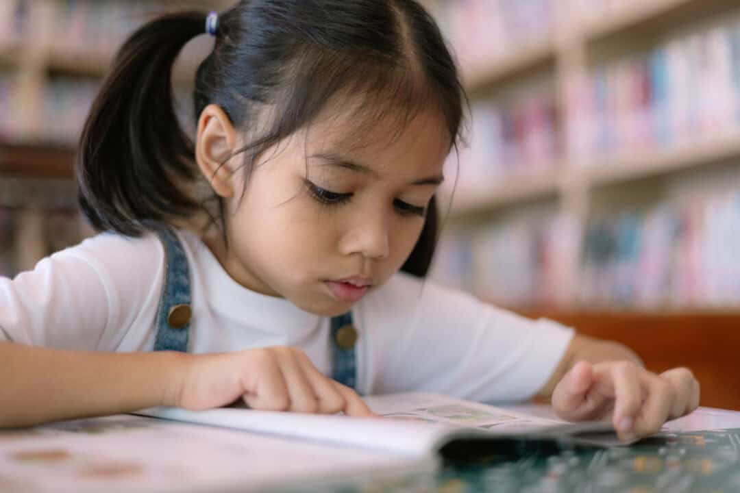A young girl is reading a money book for kids in a library. She is focused on the text and she is enjoying herself.