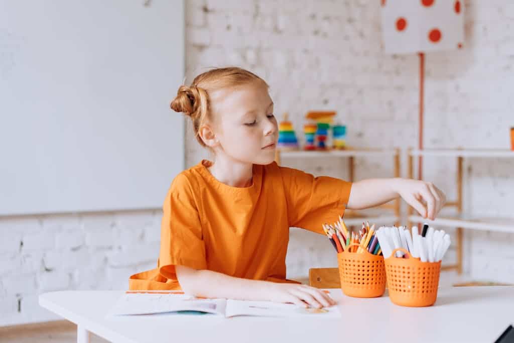 A young girl sitting at a desk with colored pencils and a cat coloring book.