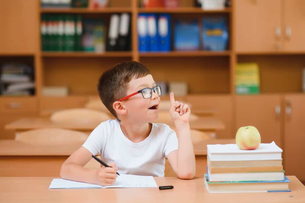 A young school boy with glasses sitting at a table with a look on his face like he just came up with a great idea. He's ready to write a sentence using different parts of speech. 