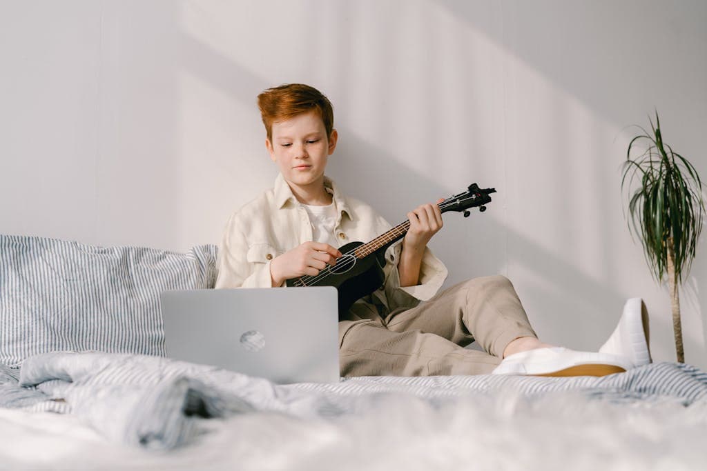 A Young Boy Sitting on His Bed while Playing Ukulele. His lap top is open as he is taking a lesson on Outschool about how to play the ukelele.
