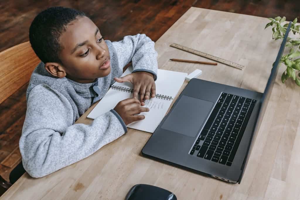 From above of smart cute African American boy studying online with netbook while sitting at table with notebook and pencil. His parents used an Outschool promo code to get $20 off his first class.