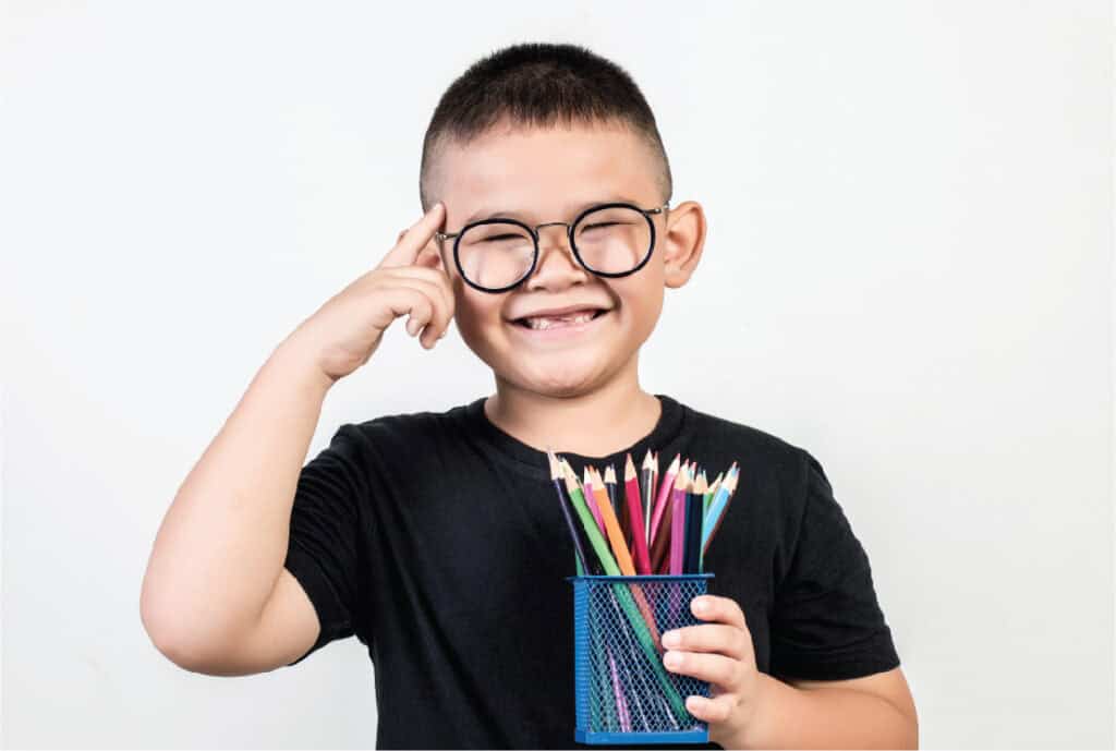 A picture of a young Asian boy with glasses holding a tin of pencils in one hand and pointing to his temple with the other hand.